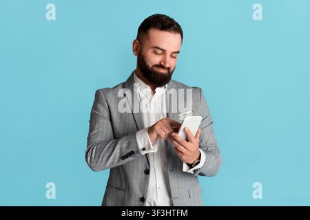 Handsome corporate employee browsing internet on smartphone, blue background Stock Photo