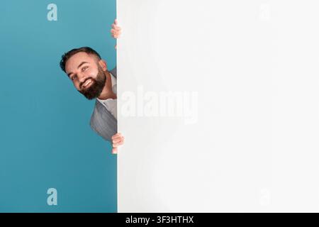 Smiling entrepreneur hiding behind blank poster with space for text or design on blue background Stock Photo