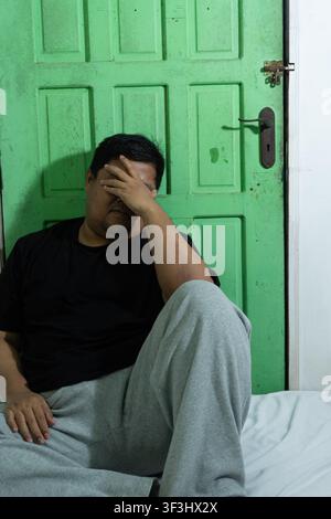 An adult Asian male sits on a white bedsheet while covering the face with one hand, expressing deep sadness, stress, and emotional exhaustion. The sol Stock Photo