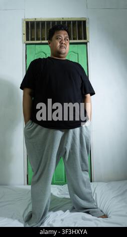 An adult Asian male stands tall on a white bedsheet with hands resting comfortably inside the pockets of grey sweatpants, looking thoughtfully toward Stock Photo
