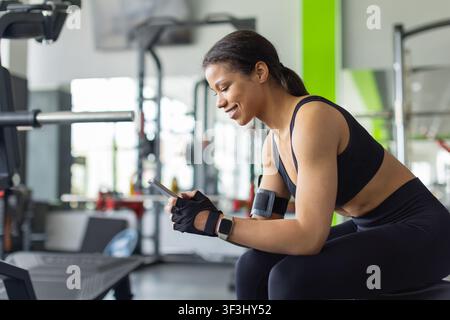 Young Black woman sitting on gym bench using smartphone during workout break Stock Photo