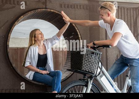 Happy young couple giving high five while meeting outdoors with e-bike. Friends greeting each other with high five in modern urban setting. Stock Photo