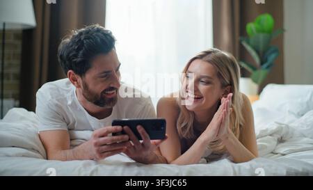 Happy Caucasian couple using mobile phone together smiling laughing lying in bed at morning. Man amazed showing smartphone to woman choosing Stock Photo