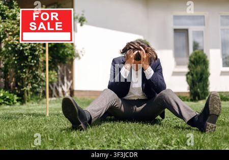 Depressed real estate agent sitting on grass in front of house for sale, outdoors Stock Photo