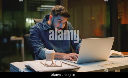 Busy multitasking Arabian business man emotional talking phone working with laptop at evening office Indian guy stressed overworked Indian businessman Stock Photo