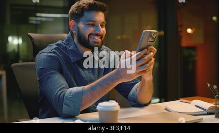 Relaxed Indian business man using mobile phone app in dark evening office smiling Arabian businessman Hindu male scrolling social media smartphone Stock Photo