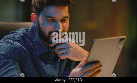 Smiling Indian business man Arabian businessman satisfied using digital tablet app at dark office at late time. Hindu male guy worker working using Stock Photo