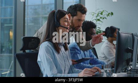 Three multiethnic business colleagues operators in call center boss coming checking work talking to woman agent. Multiracial office managers team Stock Photo