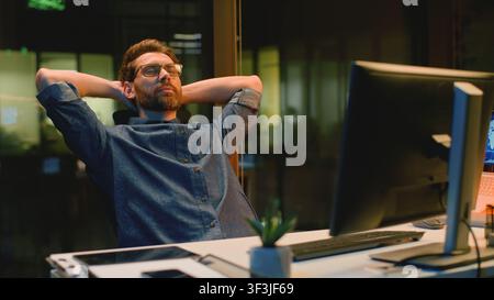 Tired overworked Caucasian business man finish online working with computer relaxing leaning at chair arms hands behind head resting businessman male Stock Photo