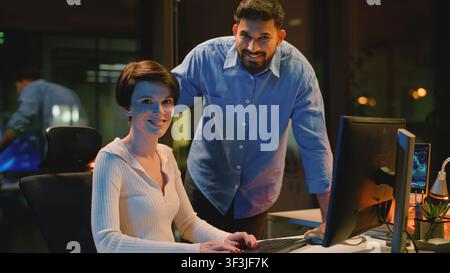 Two multiethnic business colleagues smiling looking at camera posing in dark night office. Diversity coworkers Indian businessman with Caucasian Stock Photo