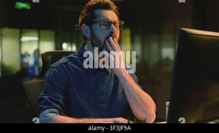 Exhausted Caucasian business man typing computer awning in night evening office overworked tired male businessman with online work yawn deadline Stock Photo