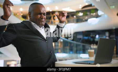 Busy African American mature business man typing laptop stretching feeling uncomfortable sitting with computer working indoors middle-aged businessman Stock Photo