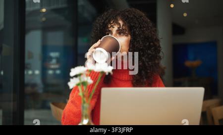 Curly haired Caucasian woman in red sweater drinking coffee from paper cup while sitting with laptop at cafe table. Female freelancer taking sip of Stock Photo