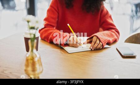 Close up of woman in red sweater writing notes with pencil in notebook at cafe table, takeaway coffee cup nearby. Female with curly hair studying or Stock Photo