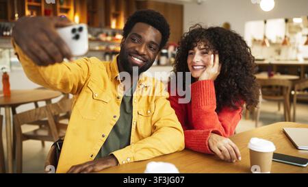 Man in yellow shirt taking selfie with smartphone while woman in red sweater laughs, sitting at cafe table. African American male and curly-haired Stock Photo