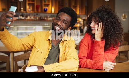 African American man taking selfie with smartphone sitting in cafe, smiling. Caucasian woman with curly hair beside him, laughing and fixing hair Stock Photo