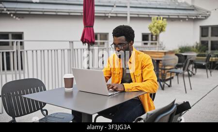 Handsome African American man with beard wearing glasses working on laptop outdoors at table. Male concentrating on computer with coffee cup beside Stock Photo