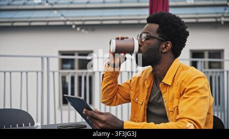 Handsome African American man with beard using digital tablet outdoors at table. Male in glasses concentrating on screen with coffee cup and notebook Stock Photo