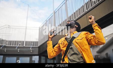 Handsome African American man with beard wearing glasses holding smartphone outdoors, looking surprised. Male reacting emotionally to good news or Stock Photo