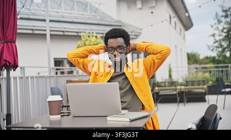 Handsome African American man raising hands in surprise while working on laptop outdoors. Male sitting at table with coffee cup and notebook, reacting Stock Photo