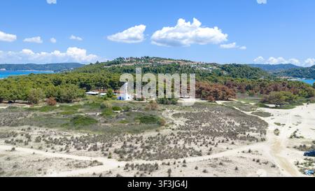 Possidi beach, Greece 11 July 2026. A white lighthouse stands amid dense green trees near a sandy beach with scattered vegetation and distant hills un Stock Photo
