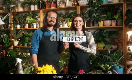 Portrait of two happy professional male and female florists holding open sign standing at flower shop looking at camera smiling. Beautiful young woman Stock Photo