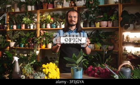 Portrait of handsome long-haired caucasian man holding and showing open sign wearing apron and glasses working at florist shop. Attractive young adult Stock Photo