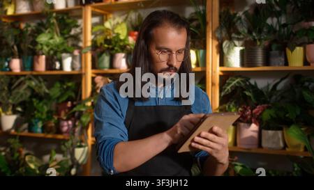 Portrait of concentrated beautiful long-haired man working as florist using tablet online typing. Handsome successful worker of flower shop working Stock Photo