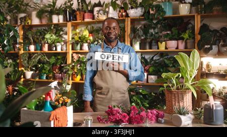 Attractive cheerful young adult African man standing at flower shop working as florist holding open sign turning to closed looking at camera smiling Stock Photo