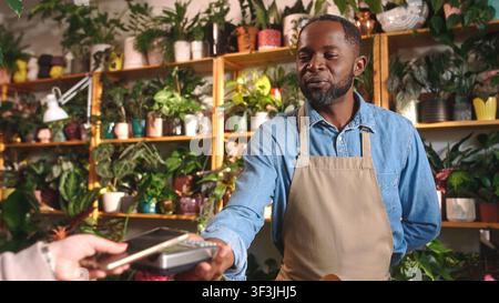 Cheerful positive attractive African American male florist selling bouquets of flowers holding payment terminal at beautiful cozy flower shop. Happy Stock Photo