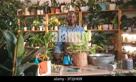 Caring concentrated African American young man taking care of flowers at florist shop using scissors wearing apron. Beautiful focused African florist Stock Photo