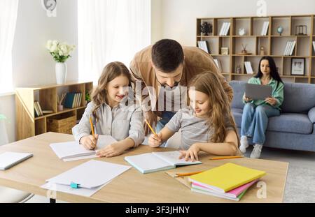 Father helping daughters with homework at home, mother working on laptop nearby Stock Photo