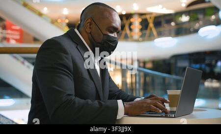 African American middle-aged business man in medical mask working with laptop in shopping mall mature businessman male professional work with computer Stock Photo