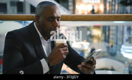 Relaxed middle-aged African American business man drinking coffee chatting mobile phone network in cafe mature businessman male user drink tea Stock Photo