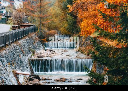 Stepped mountain stream waterfall flow through stone channel weir surrounded by vivid autumn foliage trees in seasonal alpine scenery Stock Photo
