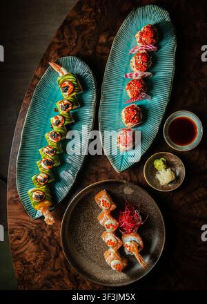Overhead professional photograph of an assortment of gourmet sushi rolls served on turquoise and dark ceramic platters with soy sauce, ginger, and was Stock Photo