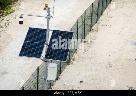 View of a solar-powered security camera pole with surveillance equipment and warning light installed along a wire fence in the rural landscape of Cala Stock Photo