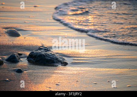 Golden hour light bathes the wet sand and sparkling water, highlighting the texture of the rippled surface and a cluster of weathered rocks Stock Photo