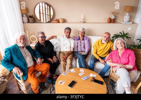 Group of diverse senior men and women sitting together in a living room, smiling and relaxing while playing a card game on a coffee table, embracing f Stock Photo