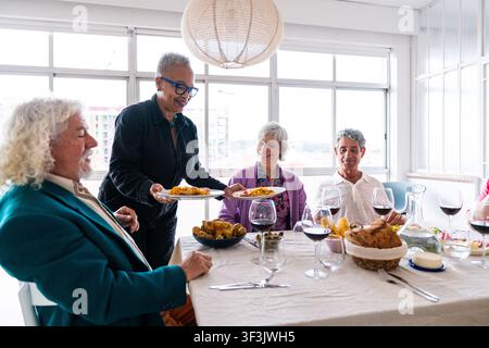 Diverse group of smiling senior adults sitting around a table, enjoying a dinner party at home, with one woman serving food, representing friendship a Stock Photo