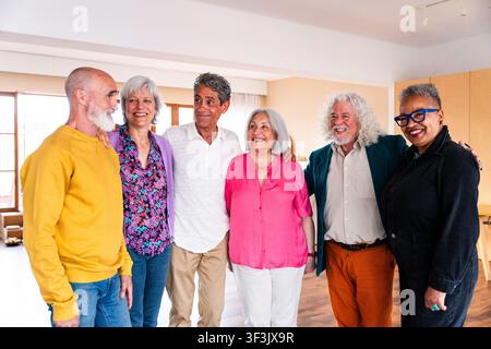 Diverse group of smiling senior adults standing together, bringing different generations and personalities in a warm, friendly indoor setting, sharing Stock Photo