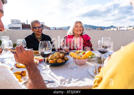 Multi-ethnic senior friends share a joyful meal with wine and food, celebrating friendship and community during an outdoor gathering on a sunny roofto Stock Photo