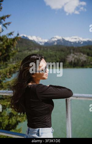 View of a young woman wearing sunglasses leaning on a metal railing overlooking a turquoise lake with snow-capped mountains in the background in Squam Stock Photo