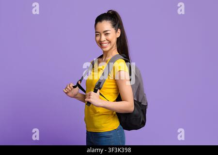 Joyful Young Asian Woman Carrying Backpack, Standing Over Purple Background In Studio Stock Photo