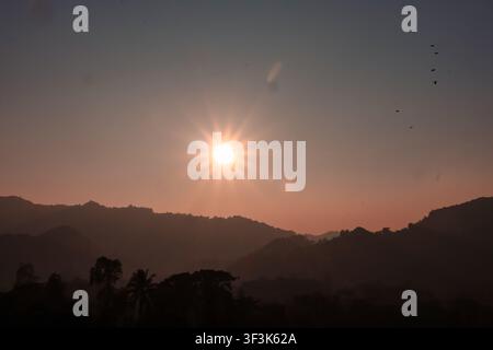View of the sun setting behind the silhouetted hills of Nhila in Teknaf, with birds flying in the sky above the landscape in Cox's Bazar, Chittagong D Stock Photo