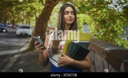 Young woman in cheer uniform holds phone and notebooks while checking screen on street; calm concentration. Stock Photo
