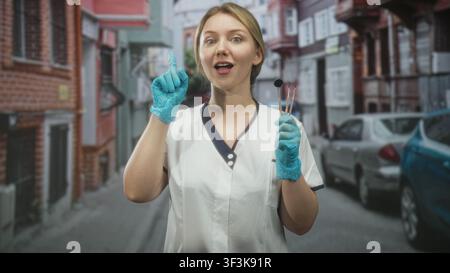 Woman dentist in uniform holding dental mirror and probes, pointing index finger while wearing blue gloves on street; reassurance care trust. Stock Photo