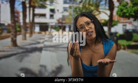 Woman in blue top beckons with both hands and open palms while tilting her head in a palm tree lined street; questioning curiosity. Stock Photo