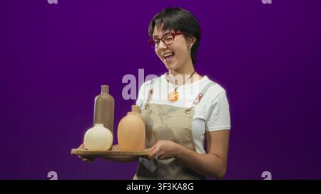Woman wearing apron holds wooden tray of three ceramic vases, smiling and presenting them with both hands in studio; creative craft joy. Stock Photo