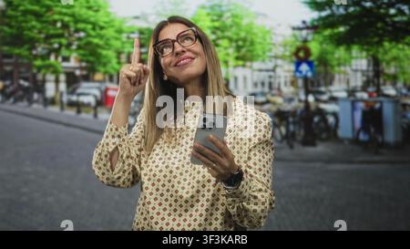 Woman holding smartphone points index finger up on a city street near bicycles and buildings; curiosity discovery inspiration. Stock Photo